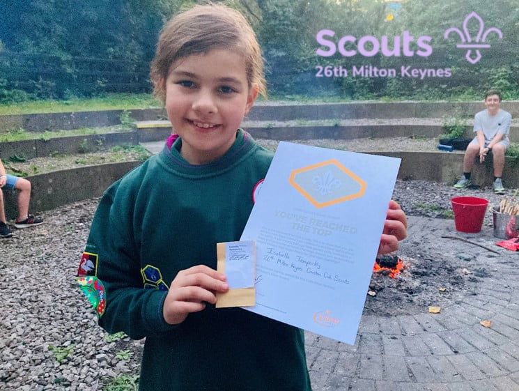 Photo of Coniston Cub Isabella wearing her green Cub Scout uniform sweatshirt, standing by a campfire and holding an envelope and certificate for her Chief Scout Silver Award.